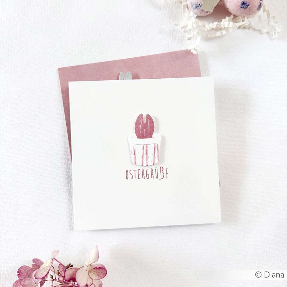 Quadratische Karte mit einem rosafarbenen Hasen in einem Muffin, hergestellt mit der Alexandra Renke Motivstanze Häschen Garten, plus "OSTERGRÜßE" über einem rosafarbenen Umschlag auf weißem Untergrund.