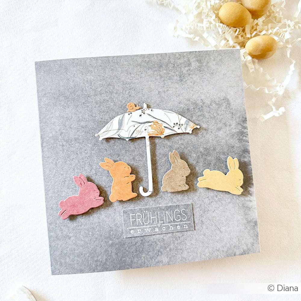 Vier pastellfarbene Hasenausschnitte von Alexandra Renke sitzen unter einem gemusterten Regenschirm auf grauem Hintergrund und tragen den Text "Frühlings erwachen".