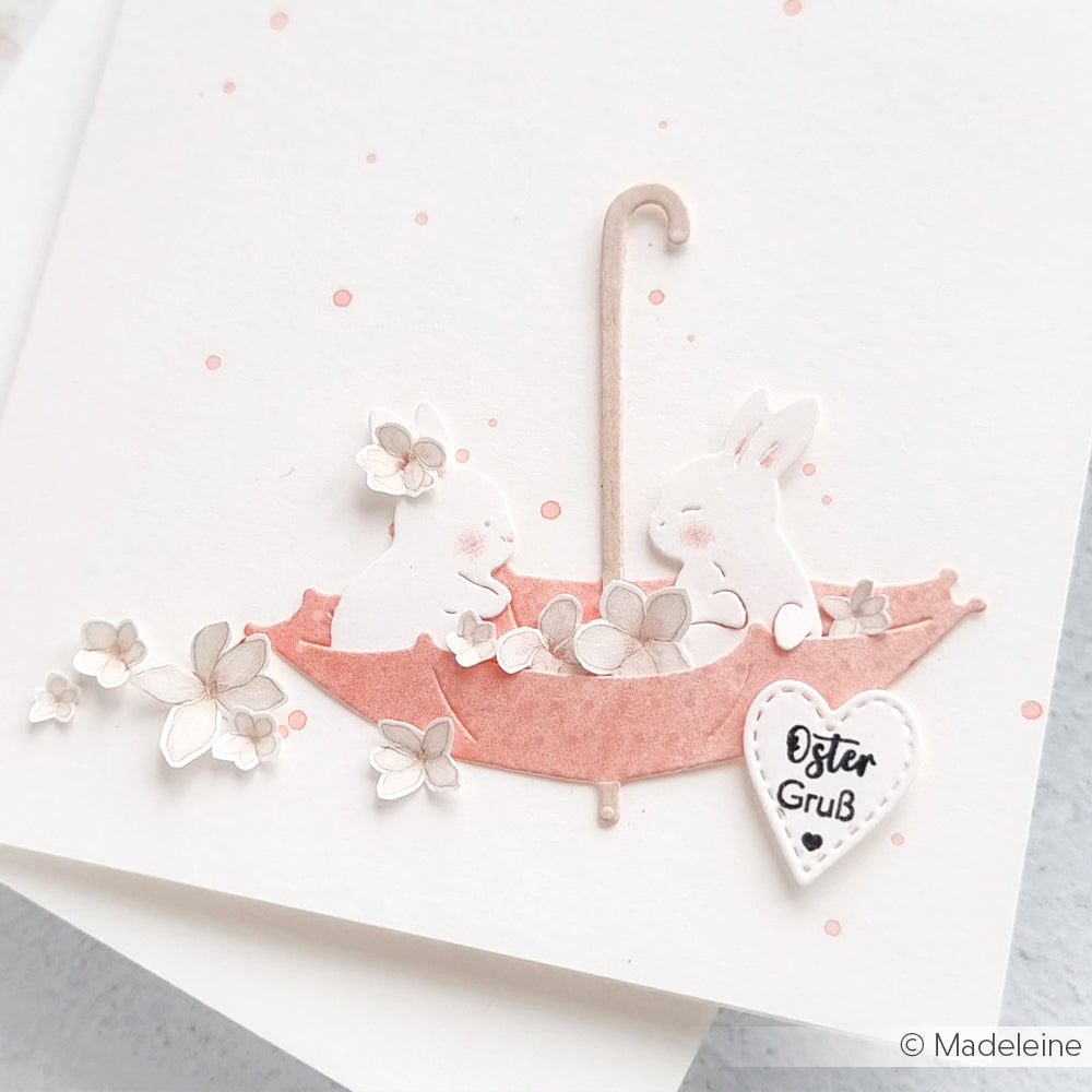 Zwei weiße Häschen sitzen in einem rosa Regenschirm mit Blumen und einem Herz mit der Aufschrift "Oster Gruß" auf einer weißen Karte, die mit Alexandra Renke Deep Edge Motivstanzen für gestochen scharfe Details hergestellt wurde.