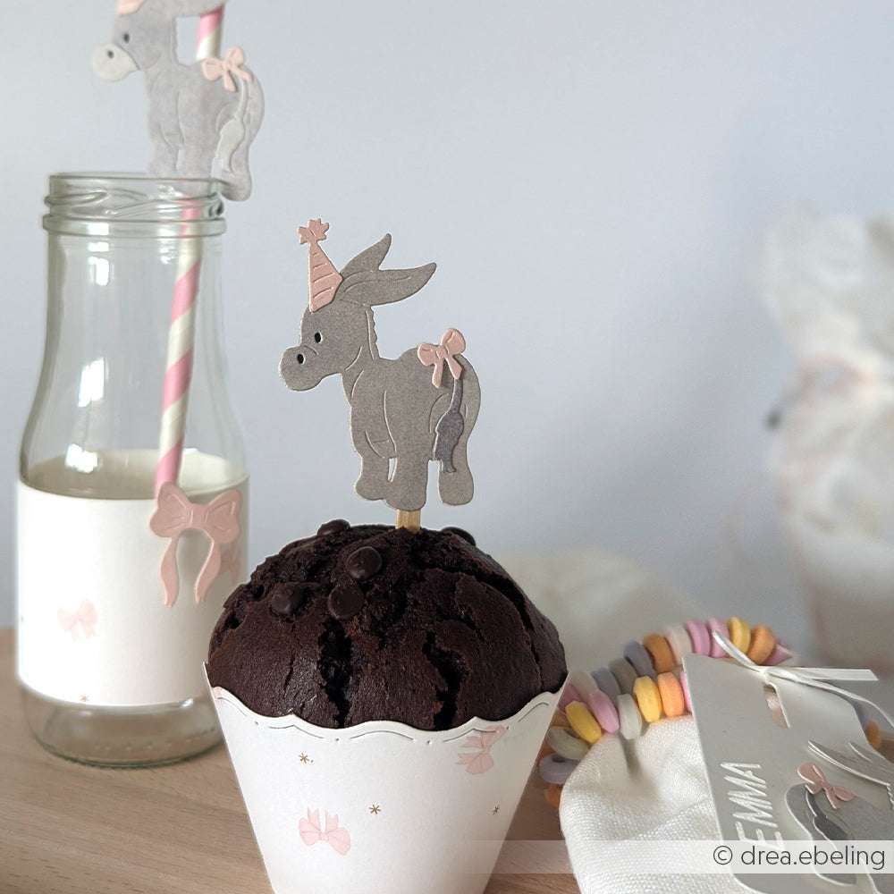 Ein Schokoladen-Cupcake mit einem Esel als Topper, der mit der Alexandra Renke Motivstanze Kuschel-Esel hergestellt wurde, sitzt neben einer Flasche und einem pastellfarbenen Bonbonarmband und verleiht dem Tisch Charme - perfekt, um den Stil von PAPPcut®ies zu unterstreichen.