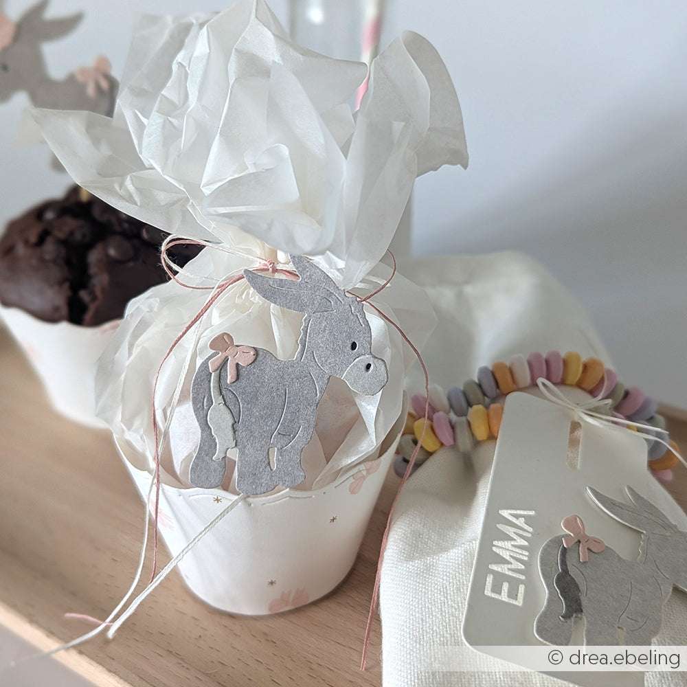 Ein in weißes Papier eingewickeltes Törtchen mit einem Eselanhänger von Alexandra Renke, daneben ein pastellfarbenes Bonbonarmband und ein Namensschild mit der Aufschrift Emma.