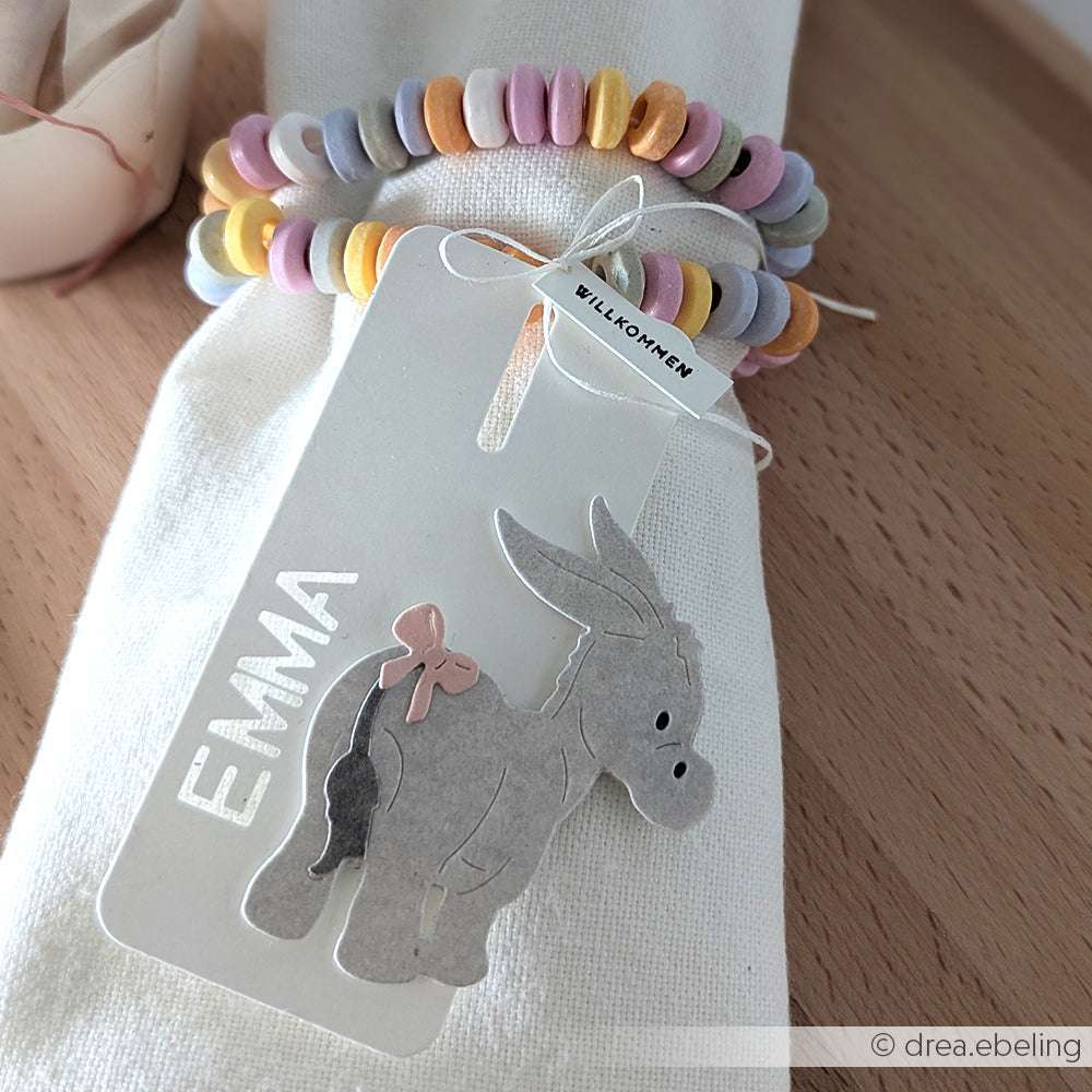 Ein Namensschild mit einer Esel-Illustration von Alexandra Renke, gefertigt mit Motivstanze, gepaart mit einem Bonbon-Armband und 'Emma' auf einer Serviette; dazu ein 'Willkommen'-Schild.