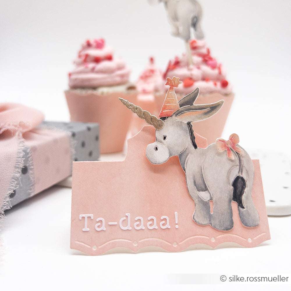 Rosa Karte mit einer Einhorn-Esel-Illustration aus der Motivstanze Kuschel-Esel, Partyhut und "Ta-daaa!"-Text. Cupcakes im Hintergrund. Ideal für jede Feier aus der charmanten PAPPcut®ies-Kollektion von Alexandra Renke.