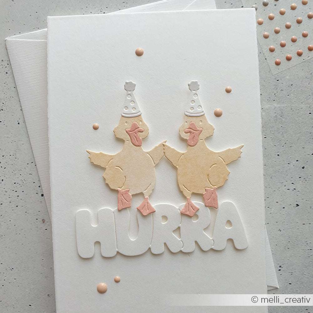 Handgemachte Karte mit zwei Partyhütchen über ausgeschnittenen Buchstaben HURRA, verziert mit pastellfarbenen Punkten und hergestellt mit Deep Edge Motivstanzen von Alexandra Renke.