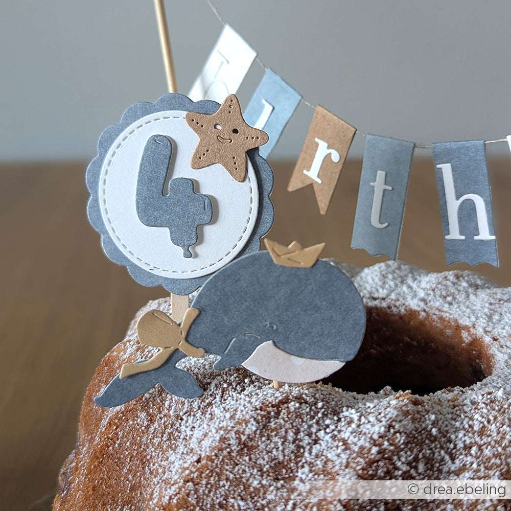Ein mit der Alexandra Renke Motivstanze Fröhlicher Wal gebastelter Papierwal und ein Sterntortenaufleger mit einer fetten "4" zieren eine gepuderte Torte; ein vierter Banner und Alexandra Renke PAPPcut®ies-Dekor bringen den Hintergrund zum Strahlen.