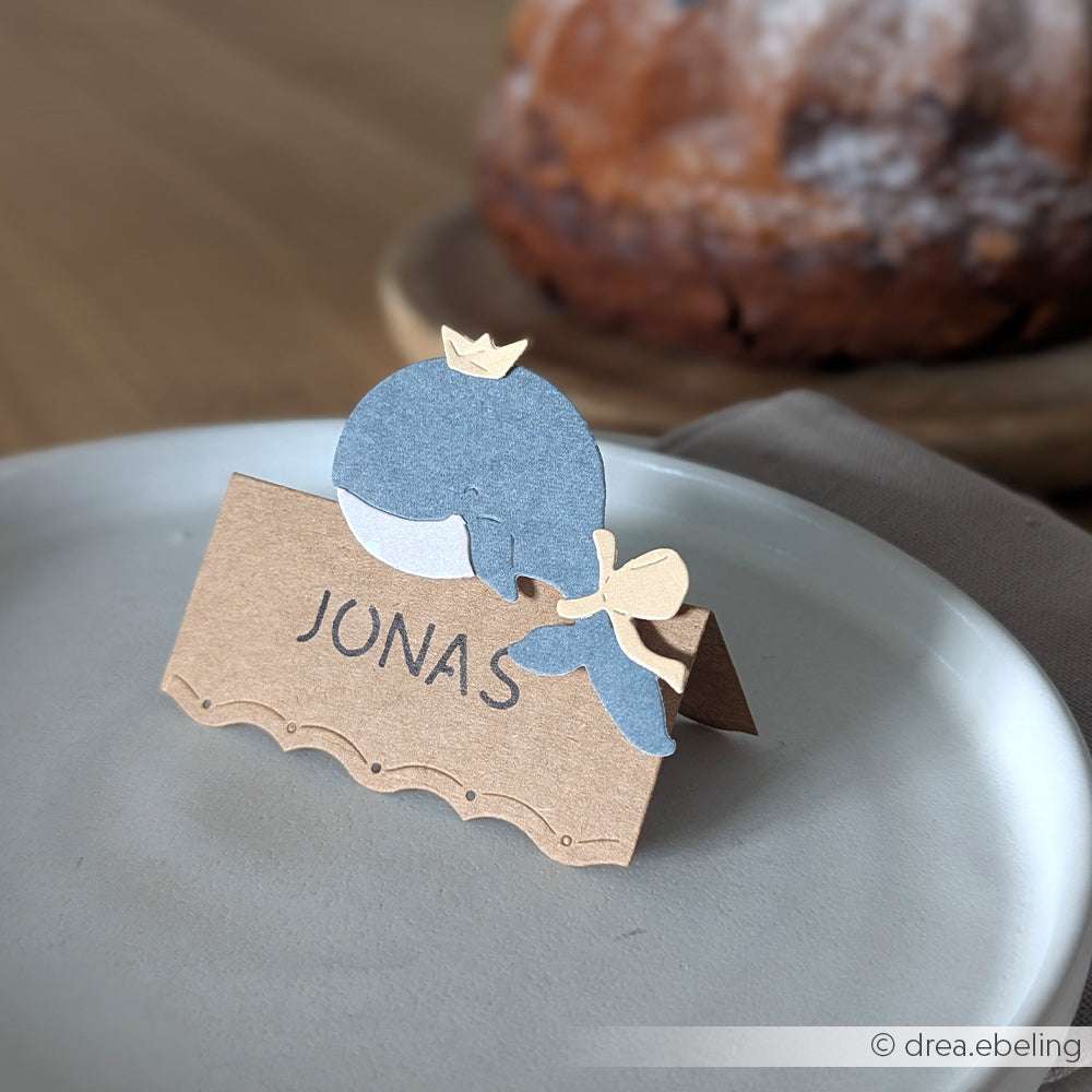 Ein Namenskärtchen aus Papier in Walform von Alexandra Renke mit der Aufschrift "Jonas" liegt auf einem Teller, der Kuchen ist im Hintergrund leicht verschwommen.