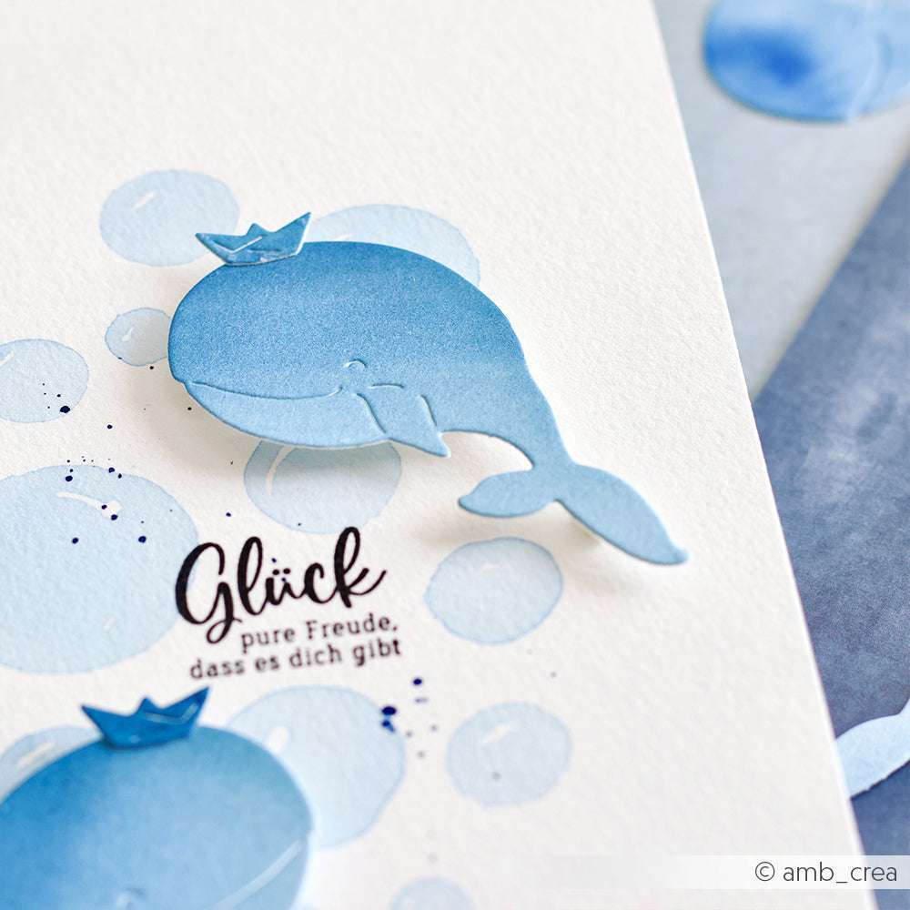 Ein blauer Papierwal mit einer Krone auf einer weißen Karte, umgeben von blauen Aquarellblasen und deutschem Text. Hergestellt mit der Alexandra Renke Deep Edge Stanze "Motivstanze Fröhlicher Wal" für zusätzlichen Charme.
