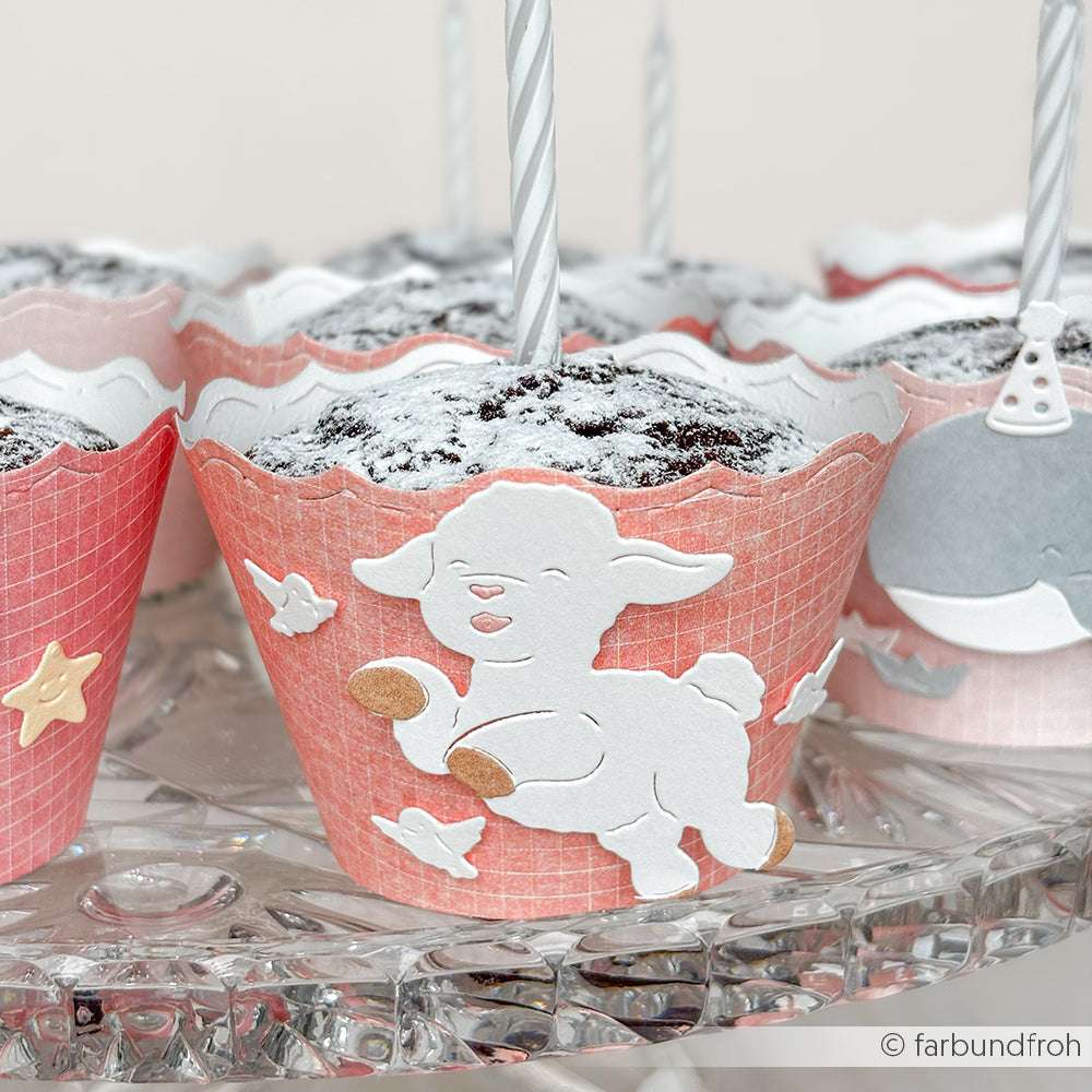 Schokoladen-Cupcakes in rosafarbener Verpackung mit weißen Lämmchen-Toppern, hergestellt mit der Alexandra Renke Springendes Lämmchen-Bowle, mit Kerzen verziert und elegant auf einem Glasständer arrangiert.