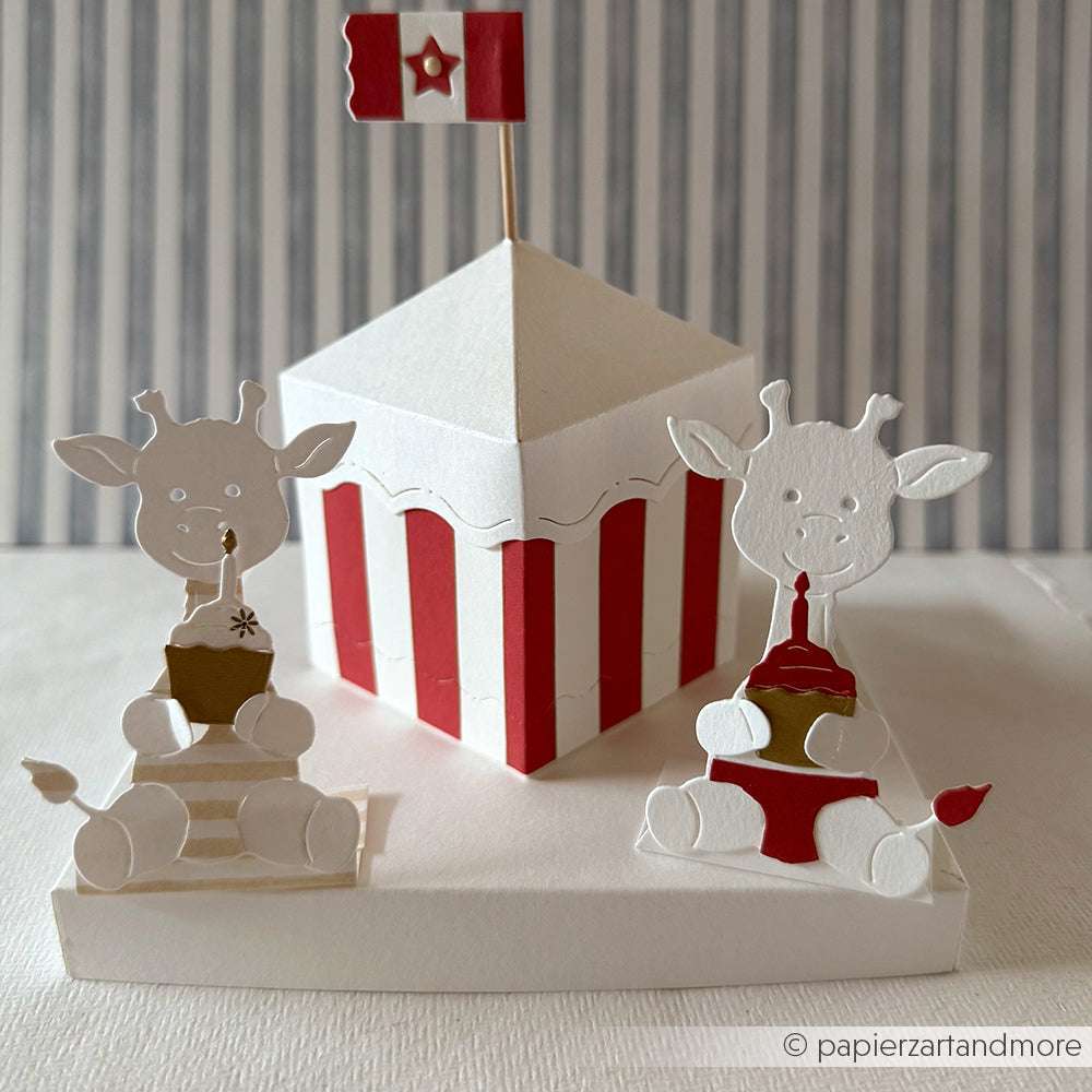 Zwei Papiergiraffen, die mit der Alexandra Renke PAPPcut Motivstanze Giraffe gebastelt wurden, tragen Partyhüte und sitzen neben einem rot-weiß gestreiften Zirkuszelt, das mit einer Flagge gekrönt ist, und bringen skurrilen Charme auf jede Feier.