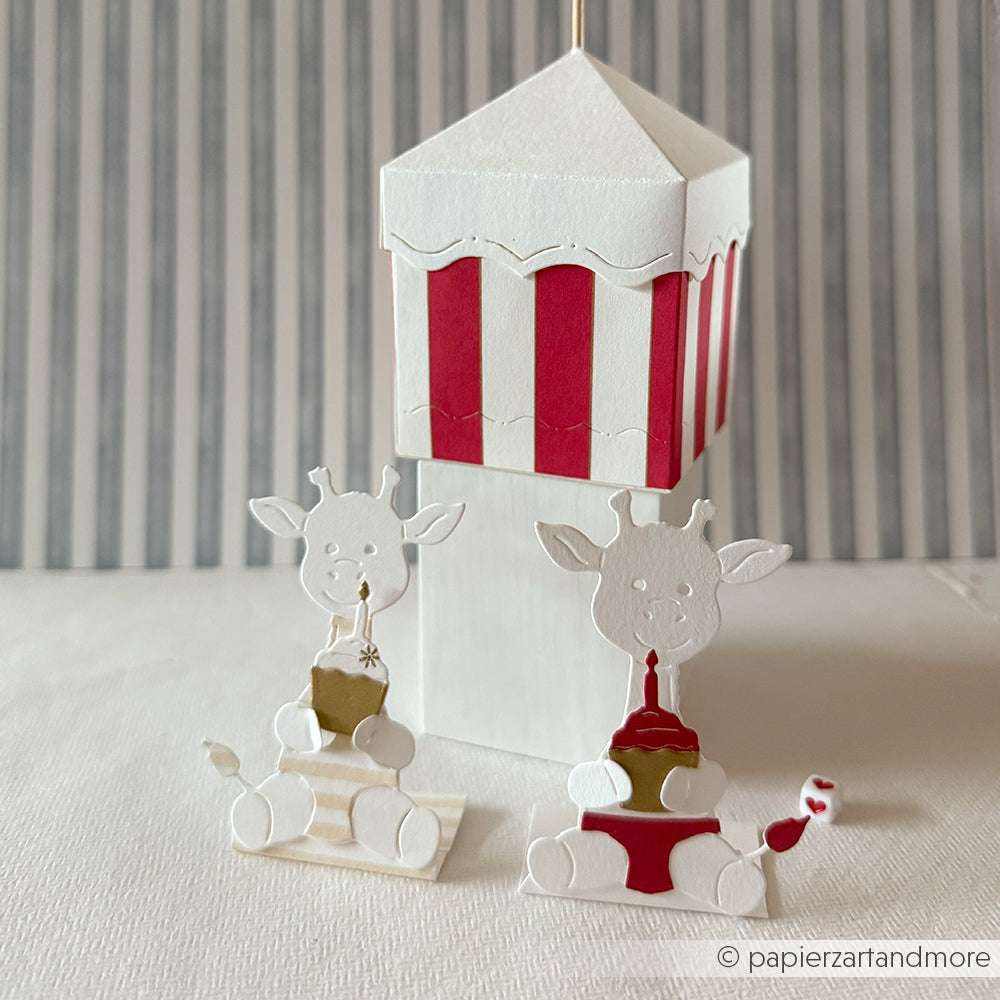 Zwei Alexandra Renke PAPPcut®ies Giraffenfiguren werden vor einer rot-weiß gestreiften Papierkarussellbox präsentiert, die jeweils mit Deep Edge Motivstanzen für präzise, detaillierte Kanten geformt wurden.