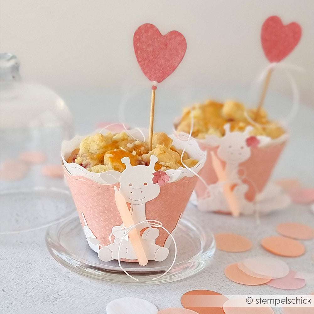Cupcake mit rosafarbener Hülle, Giraffendekoration aus der Alexandra Renke Motivstanze Süße Giraffe und Herz-Topper auf einer Glasplatte mit pastellfarbenem Konfetti.