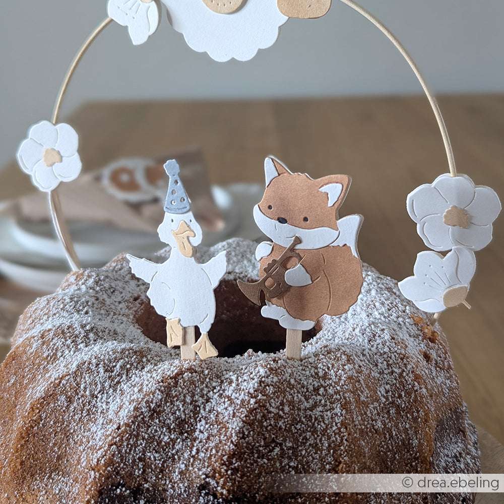 Ein Fuchs und eine Ente, die mit der Motivstanze Flauschiger Fuchs von Alexandra Renke gebastelt wurden, thronen auf einem gepuderten und mit Blumen bestückten Gugelhupf - eine bezaubernde, von PAPPcuties inspirierte Szene.