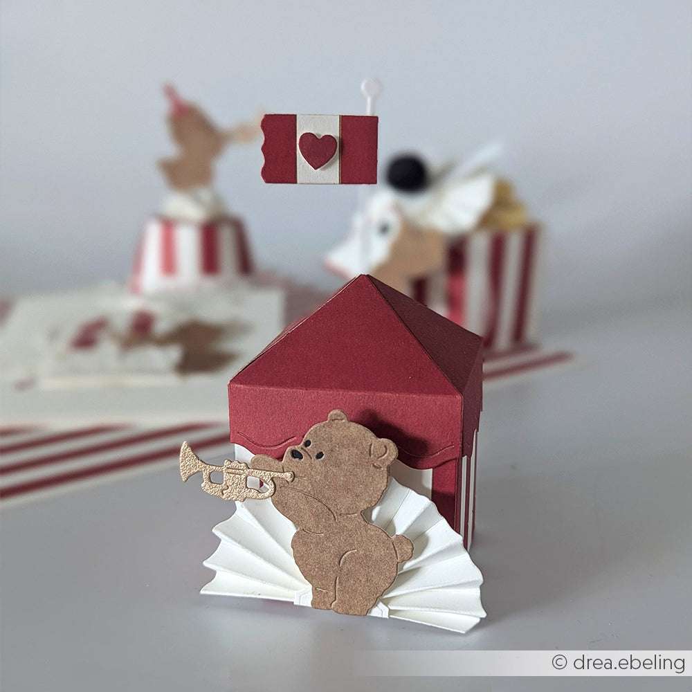 Ein mit der Motivstanze Neugieriges Bärchen von Alexandra Renke gebastelter Papierbär trompetet auf einem roten Geschenkkarton, umgeben von Herzen und Flaggenschmuck.