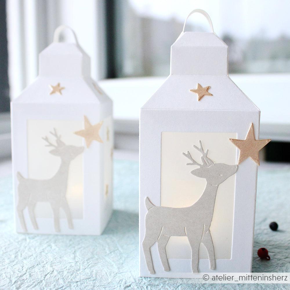 Zwei weiße Papierlaternen mit Rentier- und Sternmotiven aus der Kollektion Tierische Sternfreunde von Alexandra Renke sitzen anmutig auf einer Fensterbank.