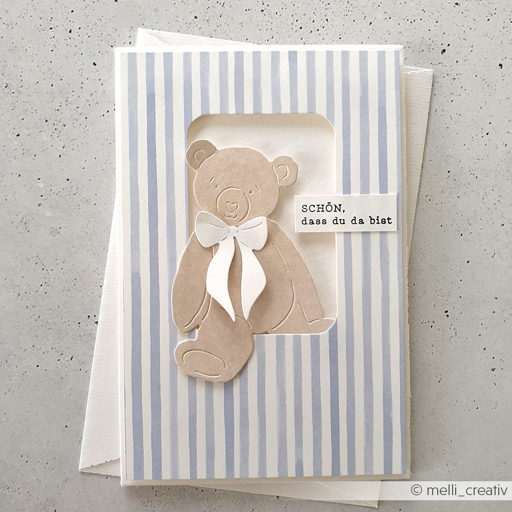 Grußkarte von Alexandra Renke mit beigem Teddybär (Motivstanze Bärchen), weißer Schleife, blauen Streifen und Text: SCHÖN, dass du da bist.