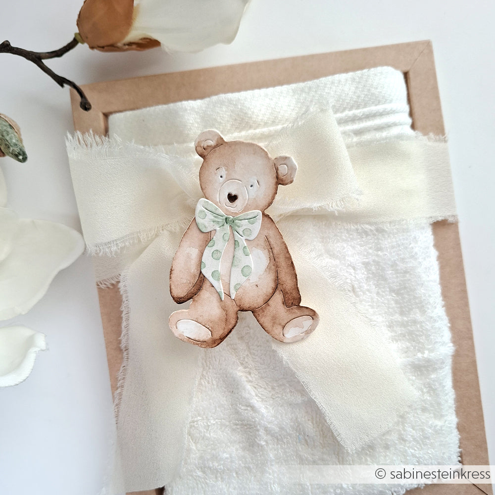 Eine Karte von Alexandra Renke mit einem aquarellierten Teddybär und einer grünen gepunkteten Schleife, gebunden mit weißem Stoffband auf strukturiertem Papier, hergestellt mit Motivstanze für zusätzlichen Charme.