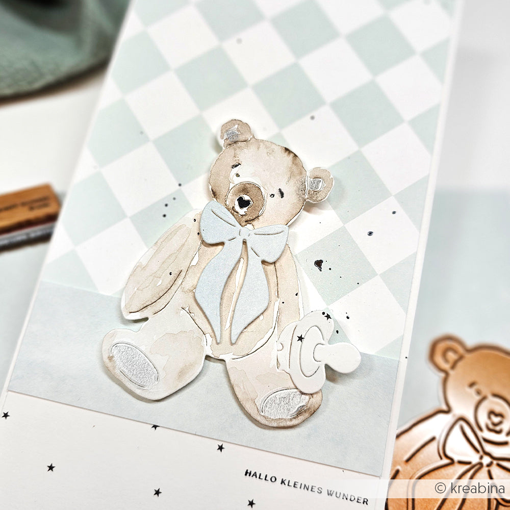Die Karte von Alexandra Renke zeigt einen aquarellierten Teddybär mit Fliege auf einem pastellfarbenen, karierten Hintergrund, der mit zarten Motivstanze-Rändern verziert ist und so für zusätzlichen Charme sorgt.