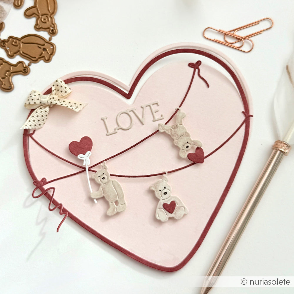 Eine herzförmige Karte mit "LOVE", drei mit der Alexandra Renke Motivstanze Kleine Bärchen Anhänger ausgeschnittene Teddybären, eine Schleife, Büroklammern und ein Stift sind auf einer weißen Fläche angeordnet.