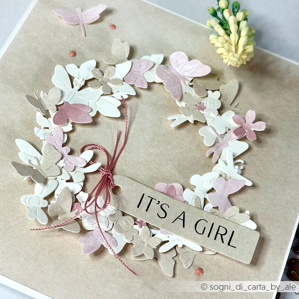 Eine Karte mit Schmetterlingskranz von Alexandra Renke, die mit Designpapier gestaltet wurde und ein Schild "IT'S A GIRL", eine rosa Schleife und pastellfarbene Blumen auf einem beigen Hintergrund enthält.
