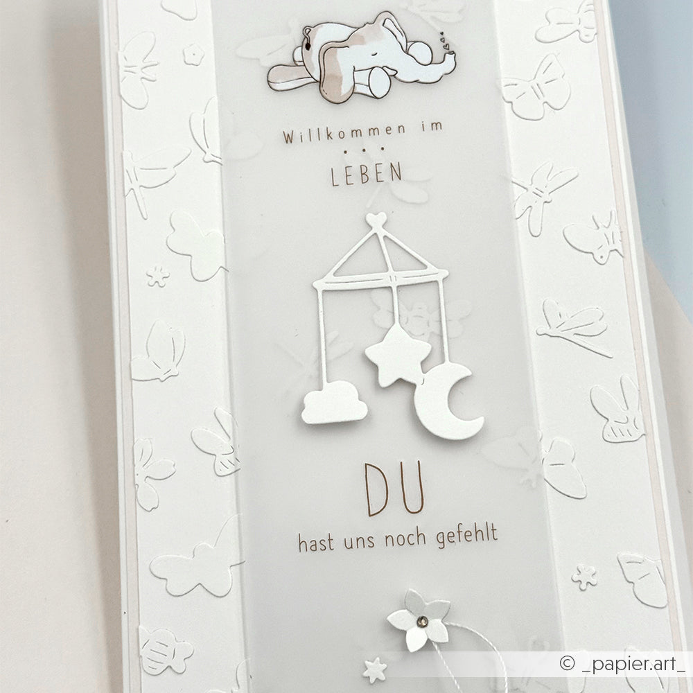 Weiße Karte von Alexandra Renke mit einem Baby, einem Handy und einem deutschen Text: "Willkommen im Leben, du hast uns noch gefehlt." Verziert mit gestanzten Wiesenzauber-Details - perfekt für alle Designpapiere und DieCut-Systeme.