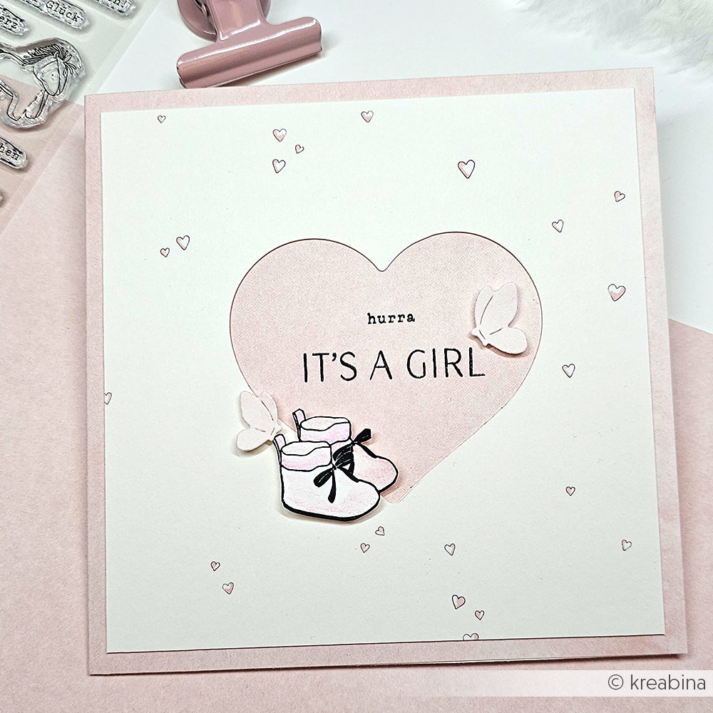 Eine rosafarbene Karte von Alexandra Renke zeigt ein Herz, Babyschuhe, Schmetterlinge und "hurra ITS A GIRL" in der Mitte, verziert mit feinen Akzenten mit Motivstanze Wiesenzauber.