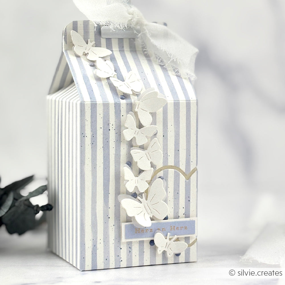 Gestreifte Geschenkschachtel, verziert mit weißen Papierschmetterlingen, die mit der Alexandra Renke Motivstanze Wiesenzauber ausgeschnitten wurden, einer goldenen Herzkontur und einem weißen Band als Abschluss.