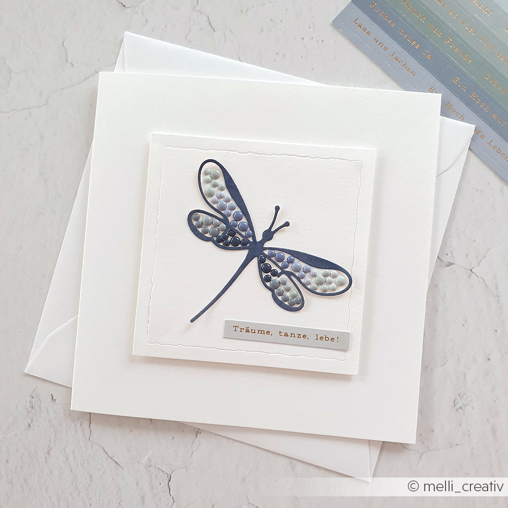 Handgefertigte Karte von Alexandra Renke, hergestellt mit Designpapier und der Lineart Libelle Stanze, mit einer blauen Libelle und dem Spruch "Träume, tanze, lebe!" auf einem sauberen weißen Hintergrund.