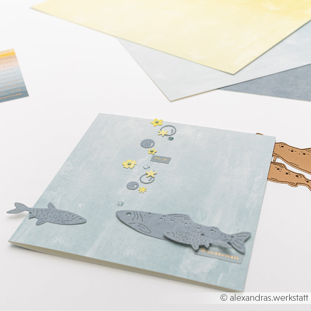 Eine handgefertigte Karte mit Fischen und Blumenausschnitten auf hellblauem Hintergrund, erstellt mit Alexandra Renke Designpapieren und der Motivstanze Fisch Duo, sowie geschichtetem Pastellpapier.