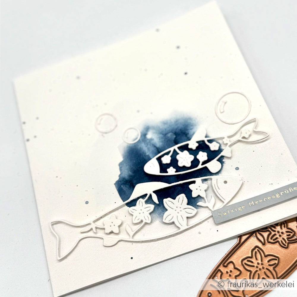 Eine Karte von Alexandra Renke mit zwei Fischen und Blumenausschnitten auf einem blauen Wasserfarbenhintergrund, verziert mit einem Metallanhänger mit der Aufschrift „Salzige Meeresgrüße“.