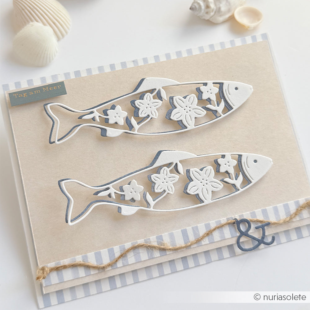 Zwei dekorative Fischausschnitte, die mit der „Motivstanze langer blumiger Fisch“ von Alexandra Renke gestaltet wurden, zieren eine Karte mit Blumenmustern, umrahmt von Muscheln und Schnur – ideal für kreative Projekte mit Stanzen für Designpapier.