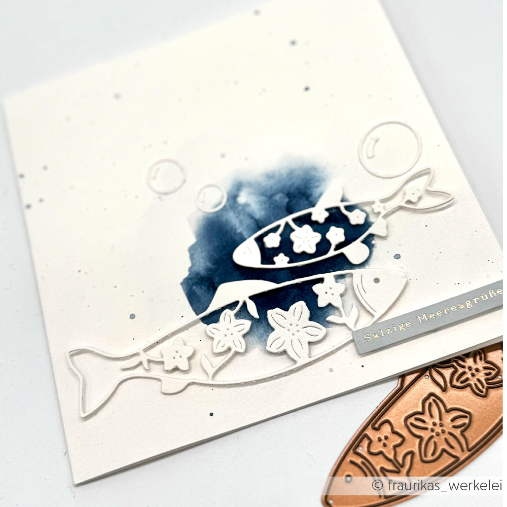 Eine weiße Karte, gestaltet mit Alexandra Renkes Motivstanze „Zwei blumige Fische“, einem blauen Aquarellhintergrund und einem metallischen Etikett mit der Aufschrift „Salzige Meeresgrüße“ – gefertigt aus hochwertigen Designpapieren und Stanzsystemen.