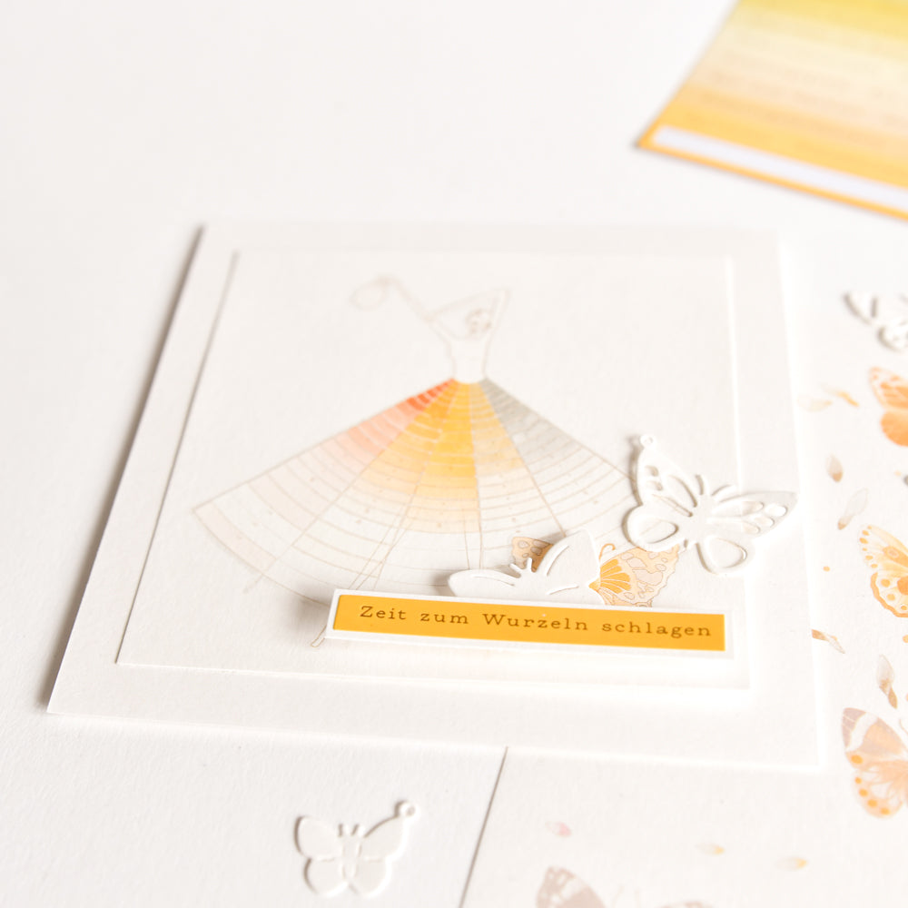 Eine zarte Karte von Alexandra Renke aus Designpapier mit einer Kleiderillustration, Schmetterlingen aus der Motivstanze Schmetterlinge und einem gelben Etikett mit der Aufschrift "Zeit zum Wurzeln schlagen".