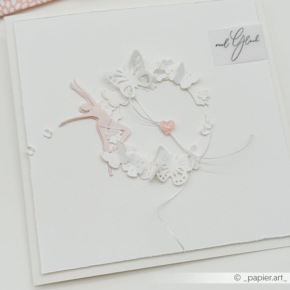Weiße Karte mit zarten Papierausschnitten, die mit der „Motivstanze Schmetterlinge“ von Alexandra Renke gestaltet wurden und Schmetterlinge, Blumen, eine Ballerina sowie ein kleines rosa Herz in der Mitte zeigen.