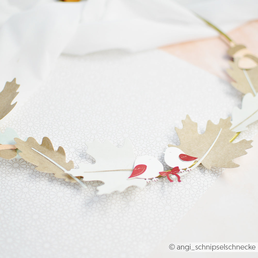 Papiervögel und -blätter, die aus erlesenen Designpapieren mit Hilfe des Motivstanzers „Vogel Duo“ hergestellt wurden, sind als zarte Girlande auf einer leicht gemusterten Fläche angeordnet.