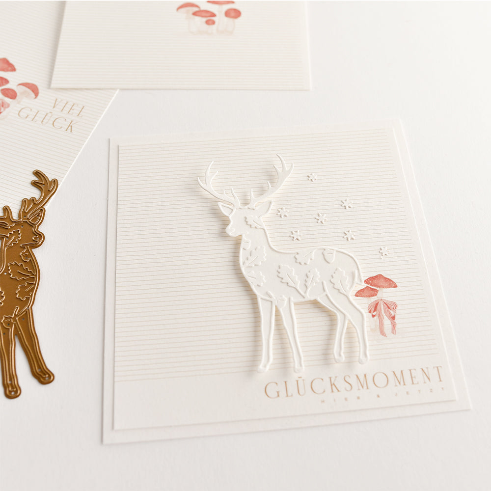 Eine quadratische Grußkarte mit einem ausgestanzten weißen Hirsch, kleinen roten Pilzen und dem Schriftzug „GLÜCKSMOMENT“, gestaltet mit einer Motivstanze im Hirsch-Design.