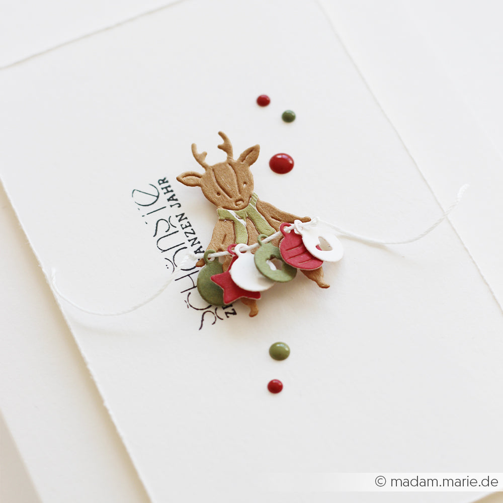 Ein aus Designpapier ausgeschnittenes Rentier, das eine Kette mit festlichen Anhängern hält.