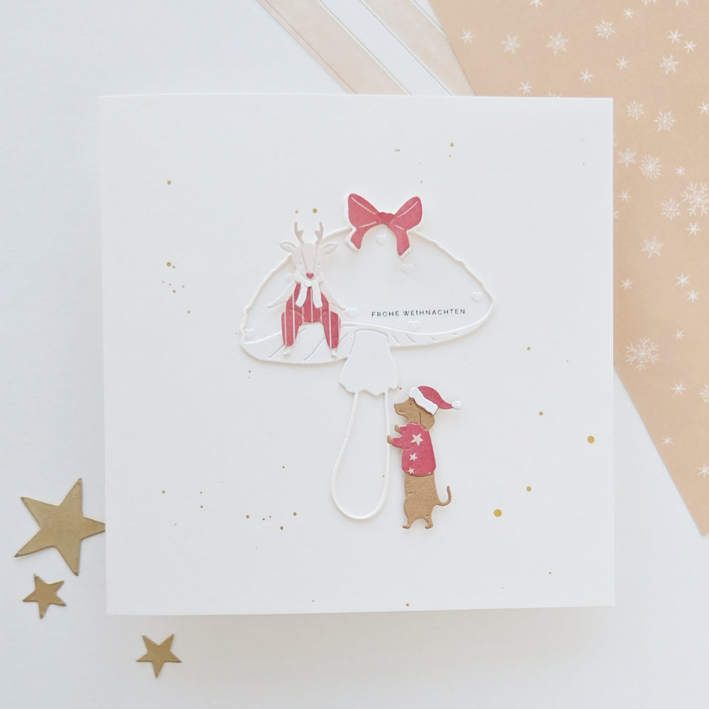 Eine quadratische Weihnachtskarte mit einem Rentier und einem Dackel, der eine Weihnachtsmütze trägt, unter einem Pilz und der Aufschrift „Frohe Weihnachten“, gestaltet mit der Motivstanze „Dackel“ und der Motivstanze „Rentier“.