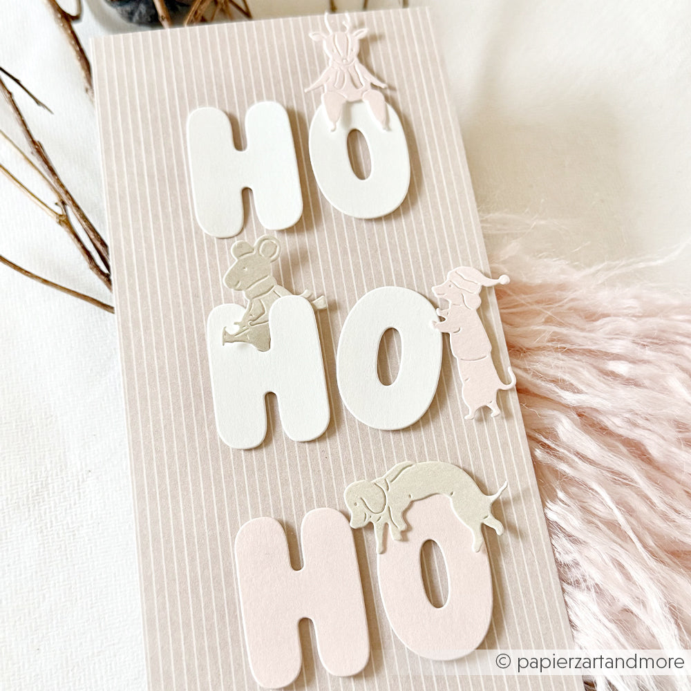 Vertikale Karte mit HO HO HO-Stanzmotiven und kleinen Tierfiguren auf Designpapier.