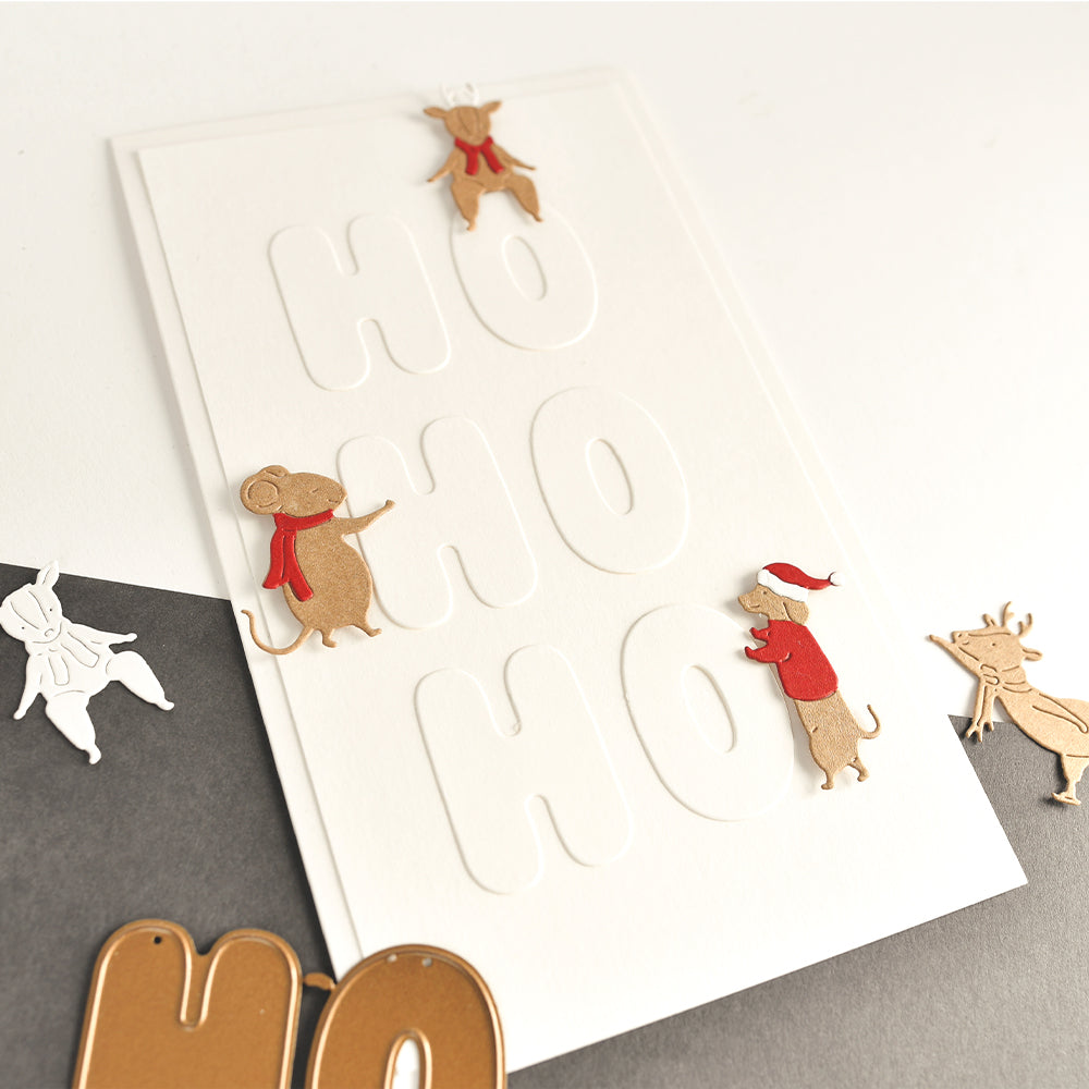 Eine weiße Karte mit der Prägung „HO HO HO“, verziert mit Motivstanz-Ausschnitten eines Rentiers, einer Maus und eines Dackels.