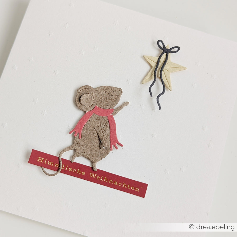 Eine Karte mit einer Papiermaus, die mit der Motivstanze „Zauberhafte Maus“ gestaltet wurde, die einen rosa Schal trägt und nach einem Stern greift, mit dem Schriftzug „Himmlische Weihnachten“.