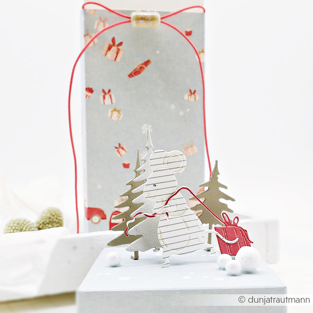 Stanzmotiv: Eine Geschenkemaus zieht eine rote Geschenkbox an zwei immergrünen Bäumen vorbei.