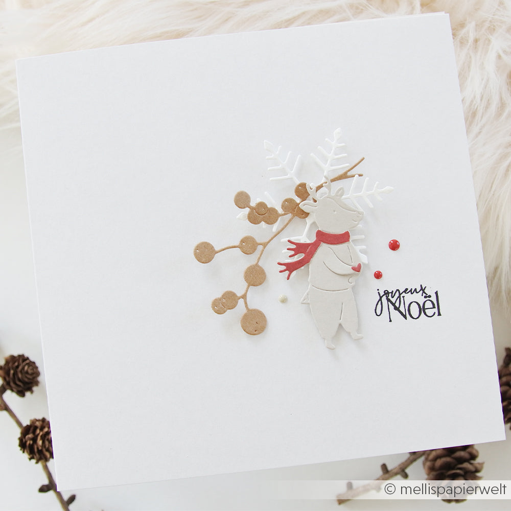 Handgefertigte quadratische Weihnachtskarte mit einem ausgestanzten Rentier, das einen roten Schal trägt, vor gemustertem Designpapier mit dem Schriftzug „Joyeux Noël“.