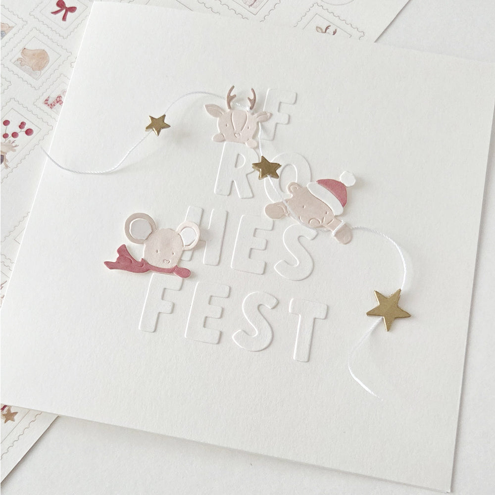 Eine quadratische Grußkarte mit dem geprägten Schriftzug „FROHES FEST“, verziert mit Tiermotiven, die mit der Motivstanze gestaltet wurden, sowie goldenen Sternen.