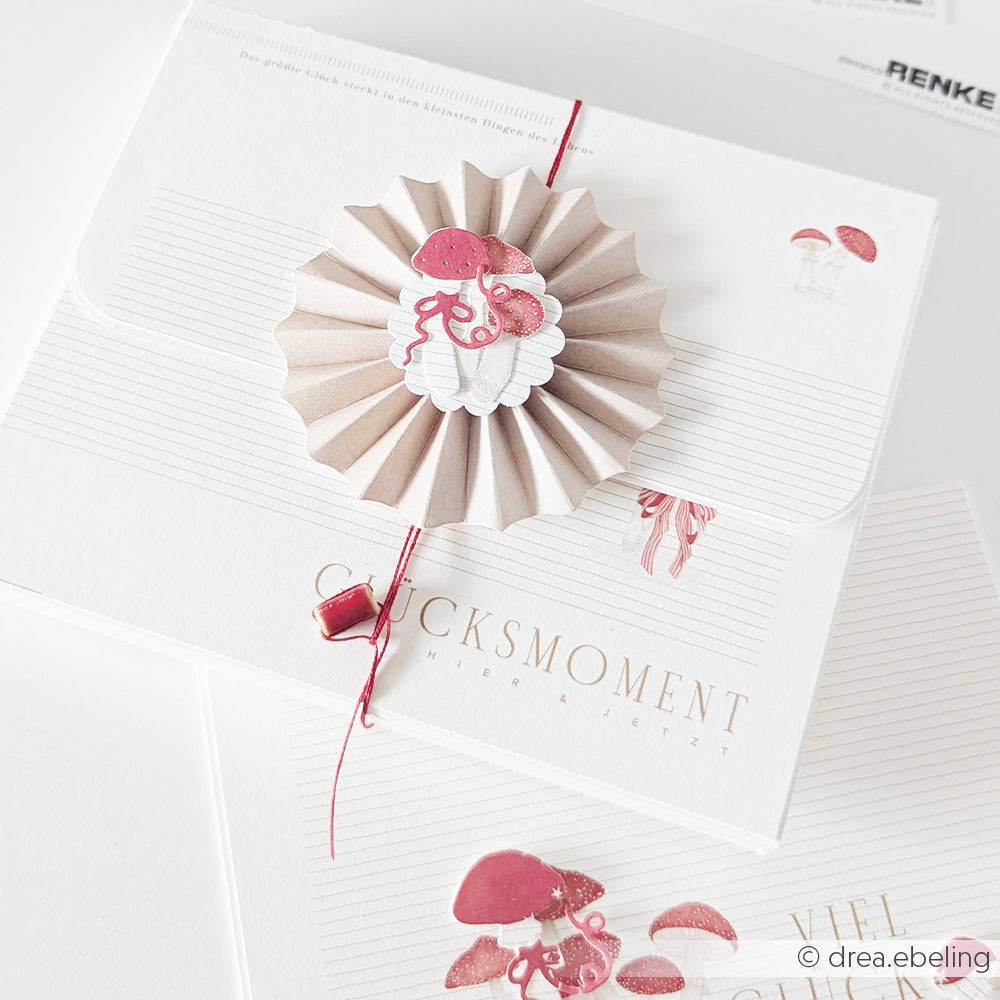 Schreibwaren-Set mit einer Papierrosette, Pilz-Illustrationen und Designpapieren im GLÜCKSMOMENT-Stil.