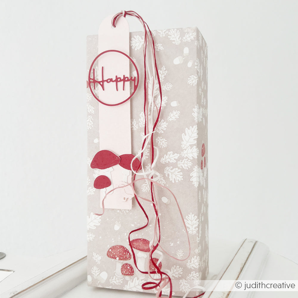 Hohe Geschenkbox, verziert mit Pilzen und einem Anhänger mit der Aufschrift Happy, gefertigt aus Designpapier.
