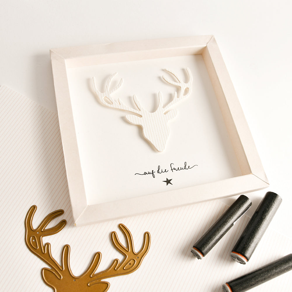 Ein gerahmter 3D-Papier-Hirschkopf mit der Aufschrift „auf die freude“ darunter, ausgestellt neben einer Metall-Hirschkopf-Motivstanze und Bastelwerkzeugen.