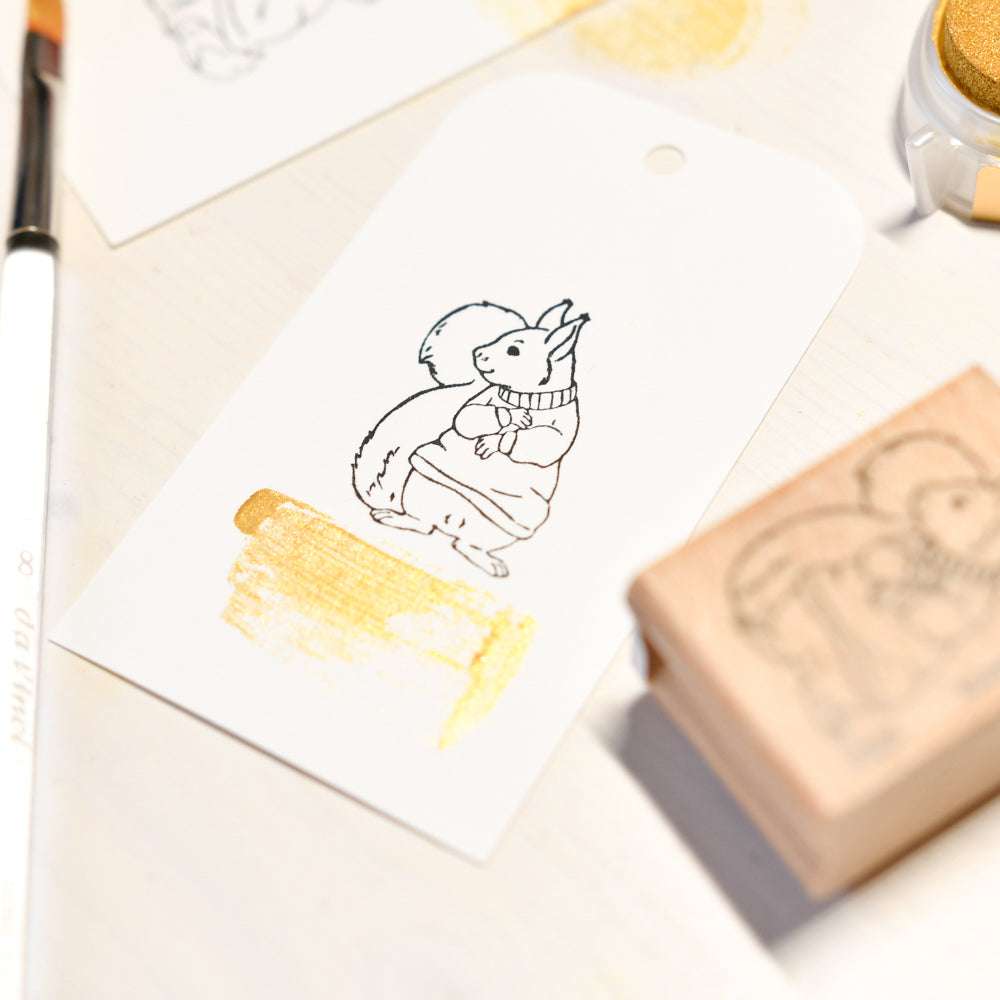 Ein gestempelter Anhänger von Alexandra Renke zeigt ein Kuschliges Eichhörnchen in einem Pullover und einem goldenen Farbmuster, daneben ein Pinsel-Ideal für Holzstempel Papierprojekte.