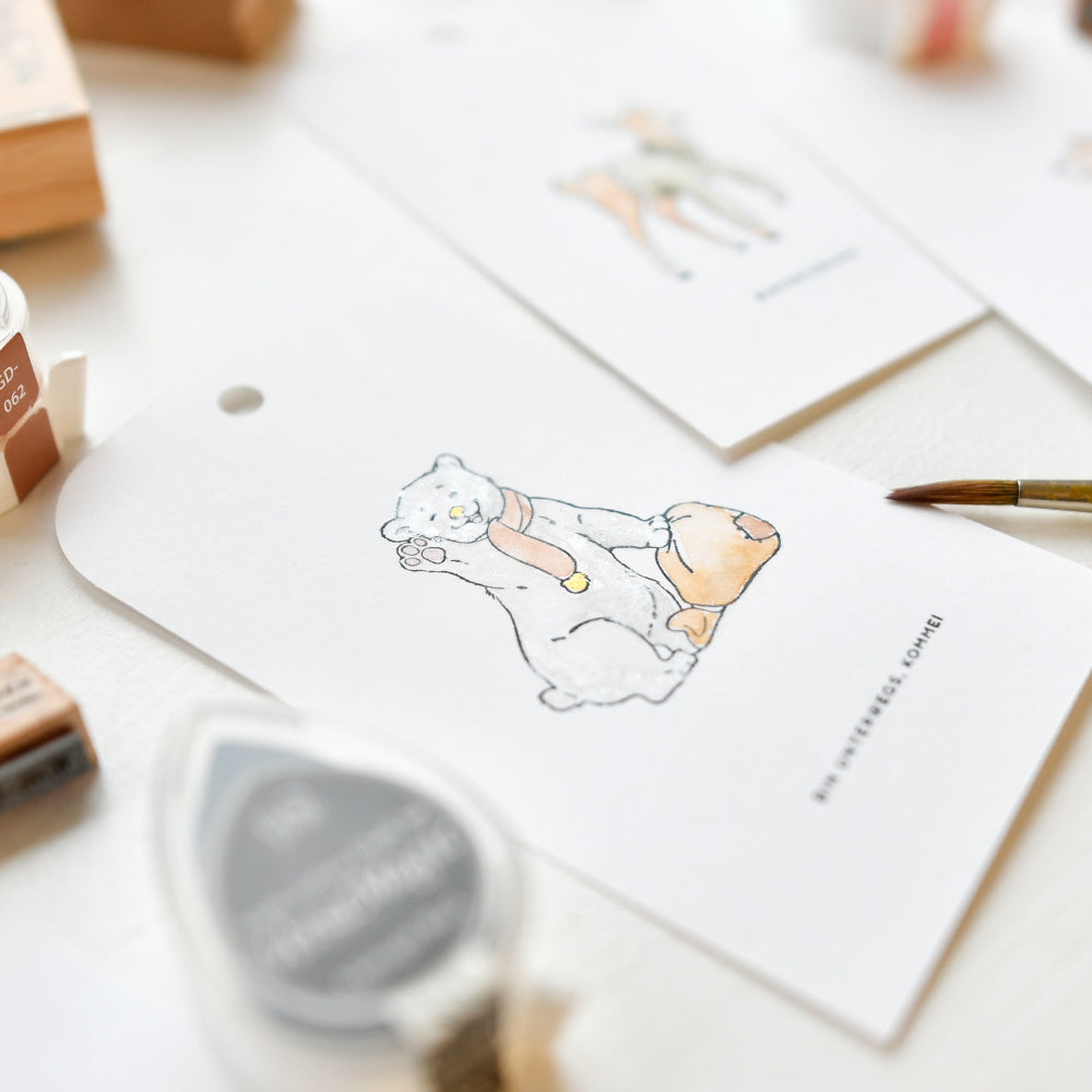 Ein handgemalter Geschenkanhänger von Alexandra Renke mit Eisbärbaby Kaja in einem Schal mit Tasche, umgeben von Malutensilien und einem charmanten Stempelmotiv.