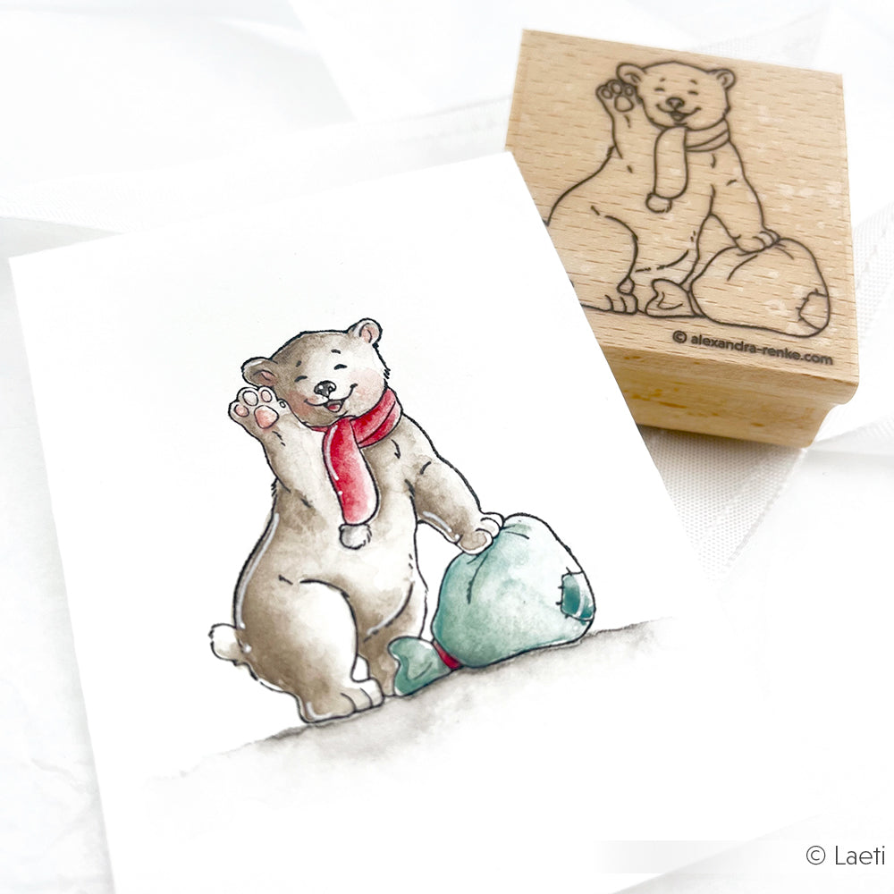 Das aquarellierte Eisbärbaby Kaja von Alexandra Renke, das einen roten Schal trägt und mit einem Fisch winkt, hat ein passendes Holzstempel-Motiv im Hintergrund.