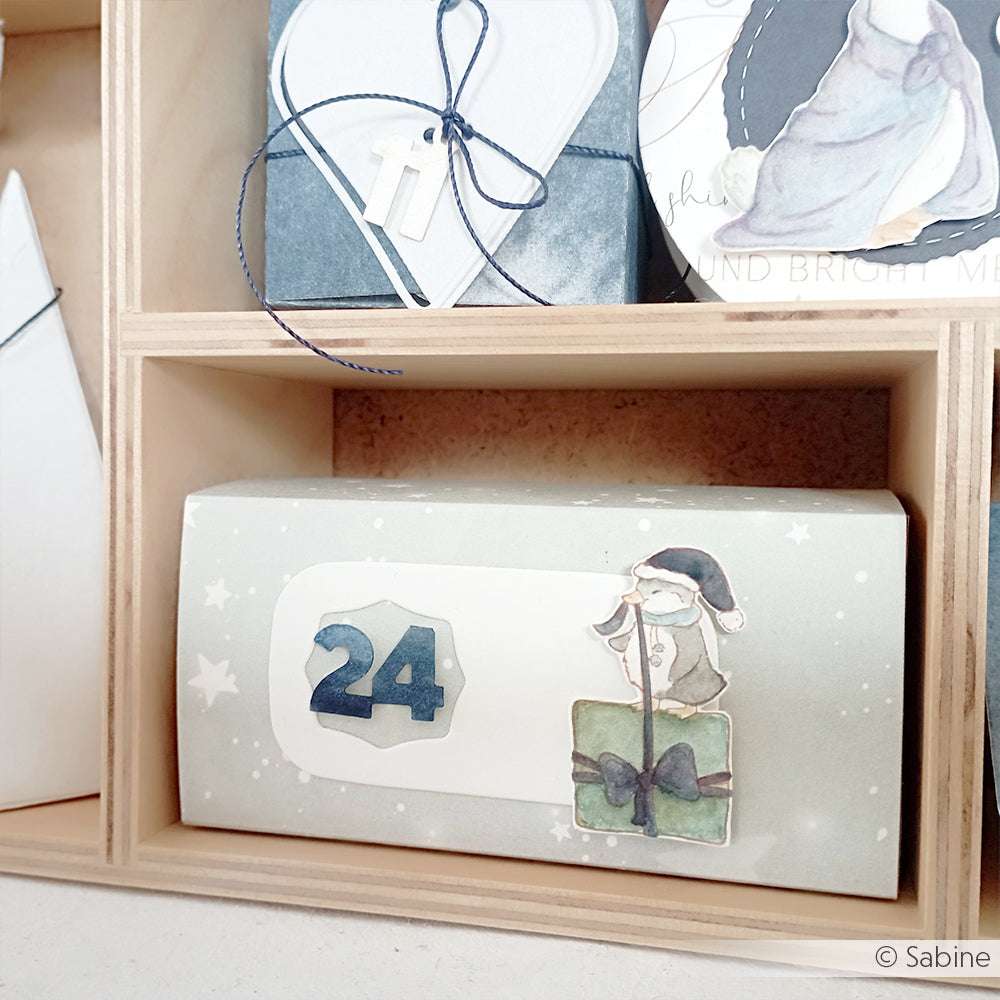 Alexandra Renke Geschenkbox Nr. 24, mit einem Mädchen mit Hut, das ein Geschenk in der Hand hält - ideal für Papierprojekte oder zur Aufwertung Ihrer Weihnachtsentlein-Sammlung - präsentiert auf einem Holzregal.